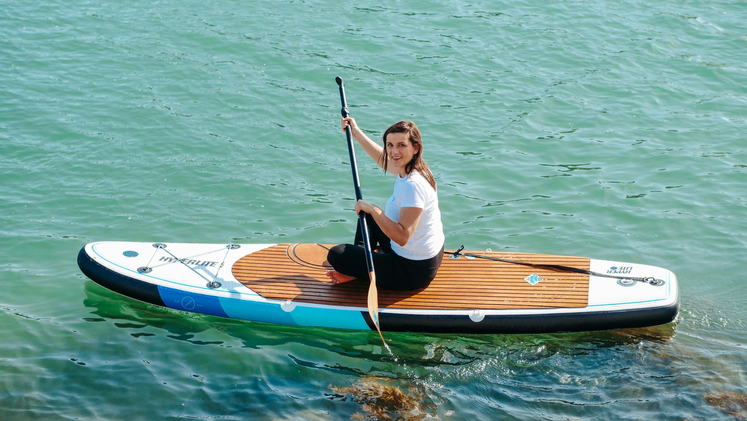 Woman sitting on paddleboard on calm water, smiling at camera.